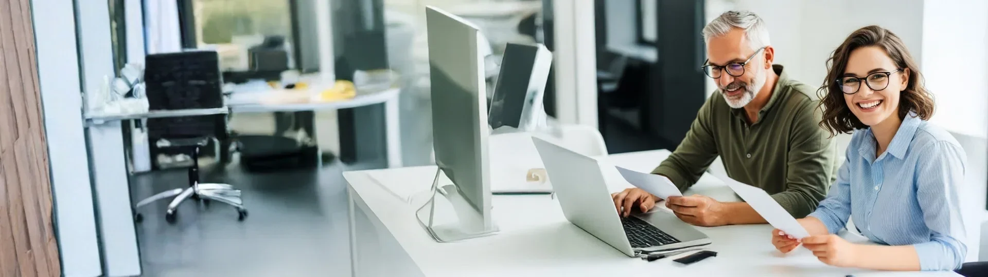 Ein Mann mit grauem Bart und eine Frau mit Brille arbeiten gemeinsam an einem Laptop mit Diagrammen und Dokumenten auf dem Tisch.